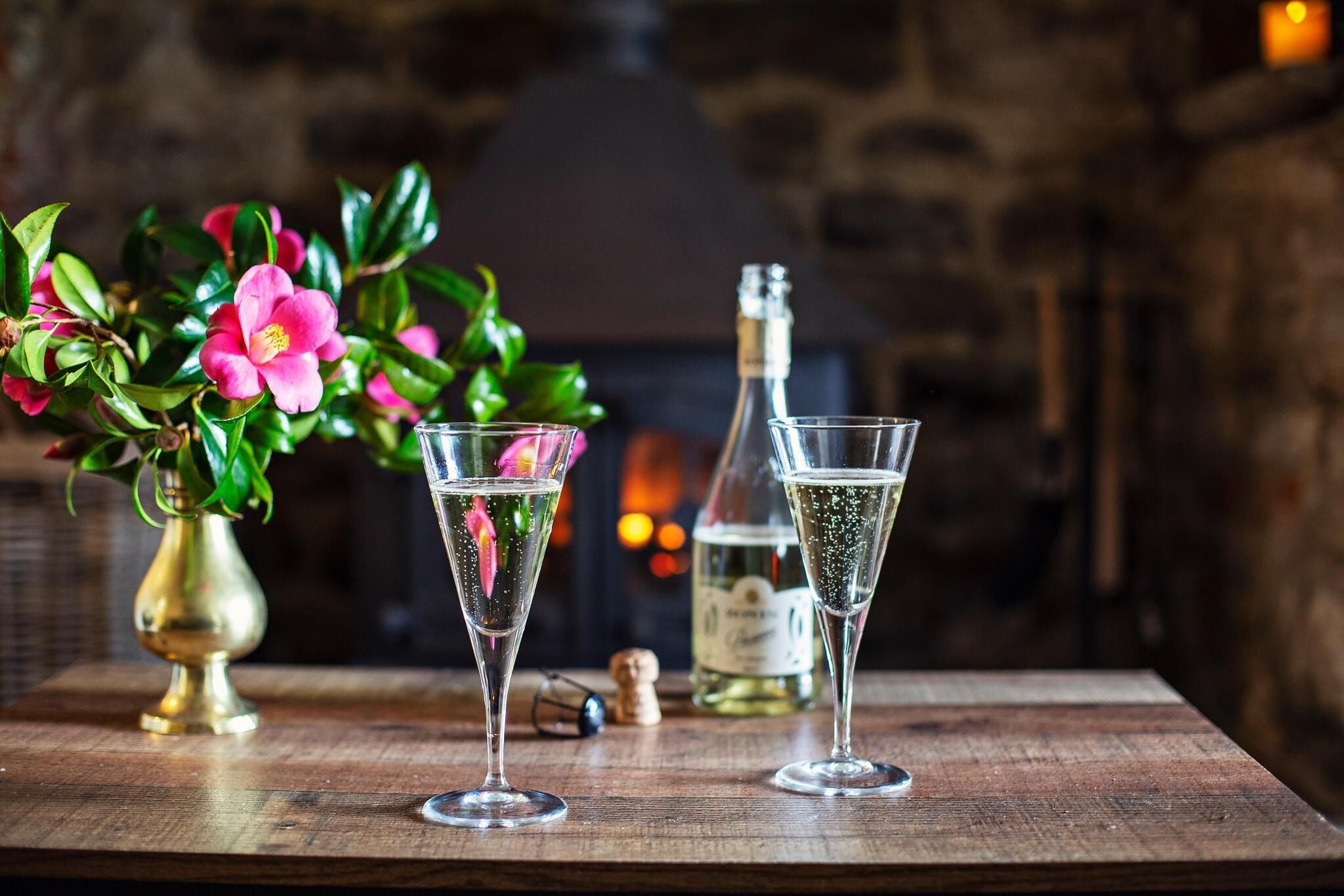 Two glasses of champagne on a wooden table with a bottle and a corkscrew, a vase with pink flowers, and a fireplace in the background.