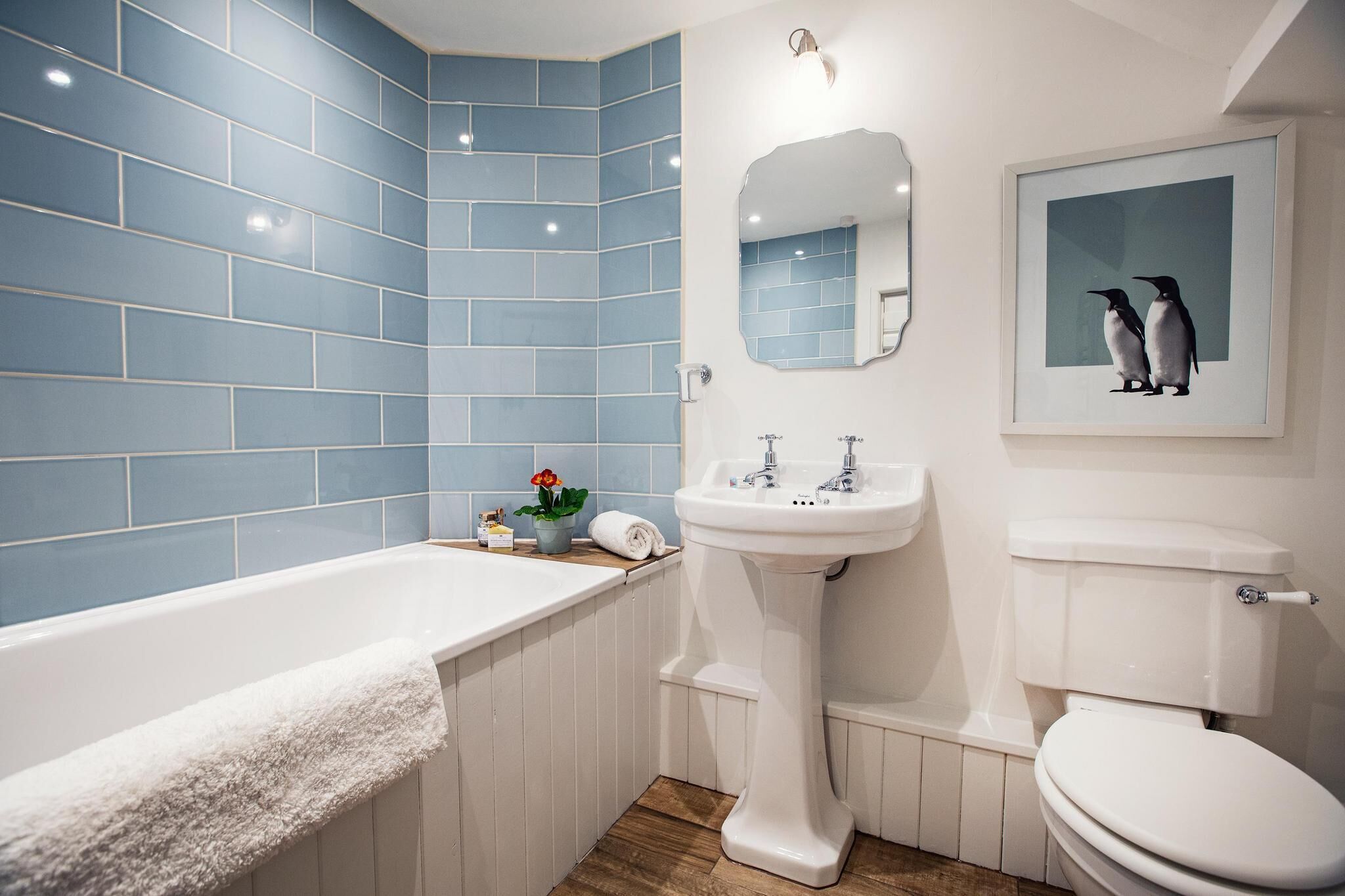 A bathroom with a white bathtub, a white sink, and a toilet. There is a mirror above the sink and a framed picture of penguins on the wall.