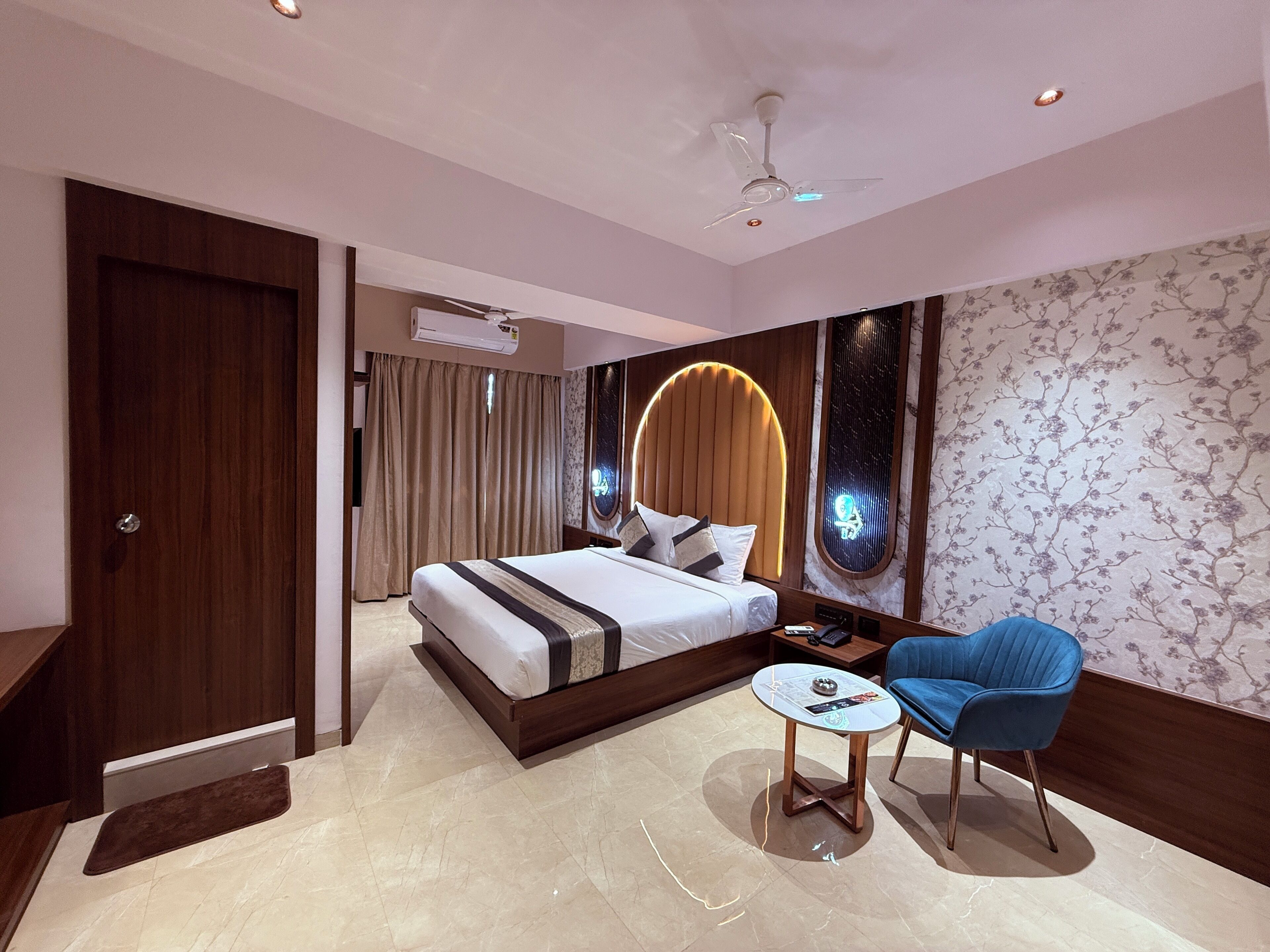 Una camera d'albergo moderna con un letto grande, un tavolino rotondo, una sedia blu e una ventola a soffitto.