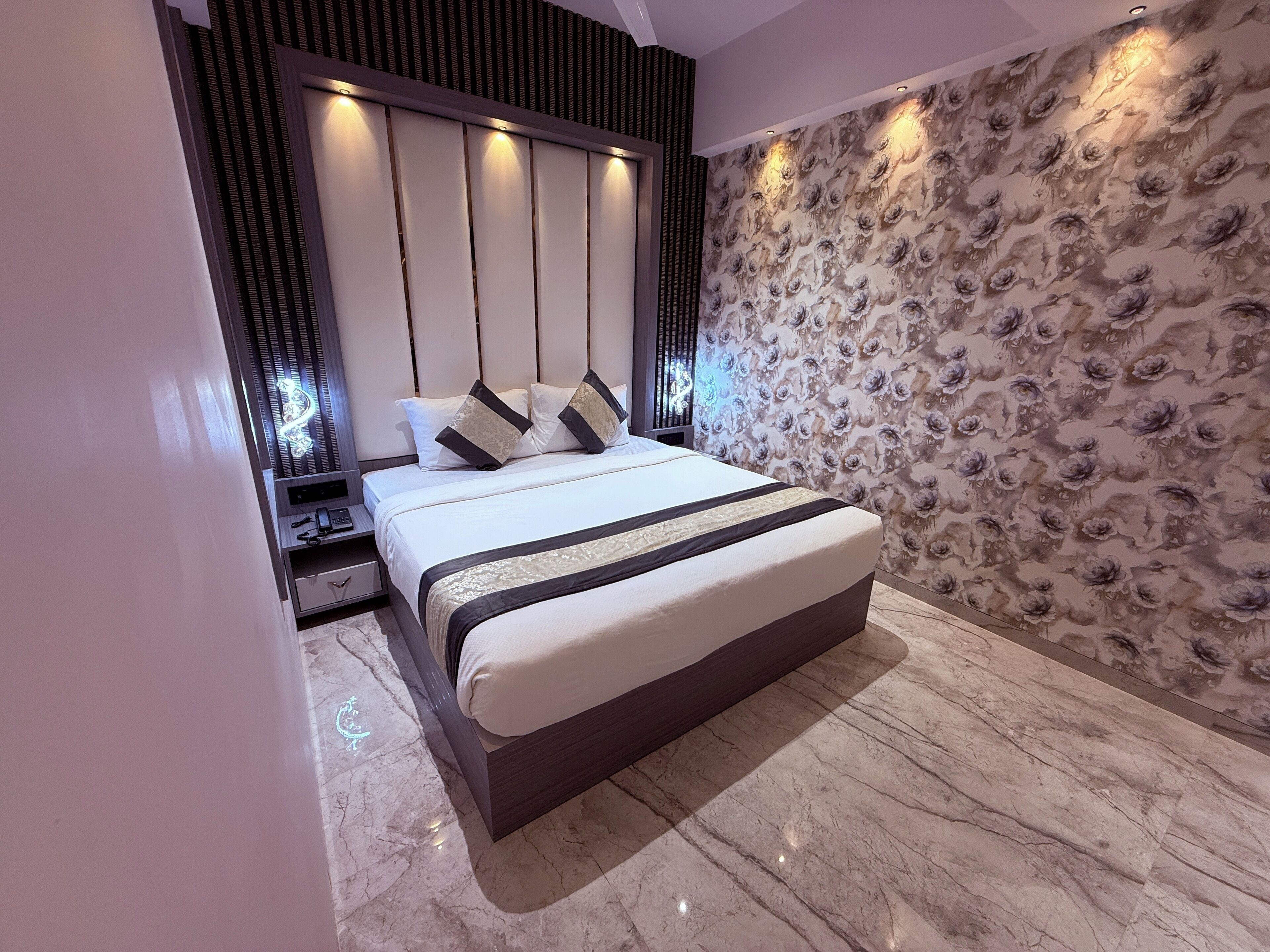 Une chambre moderne avec un grand lit, une tête de lit dotée d’un éclairage intégré, une table de chevet et un papier peint à motifs floraux sur le mur.
