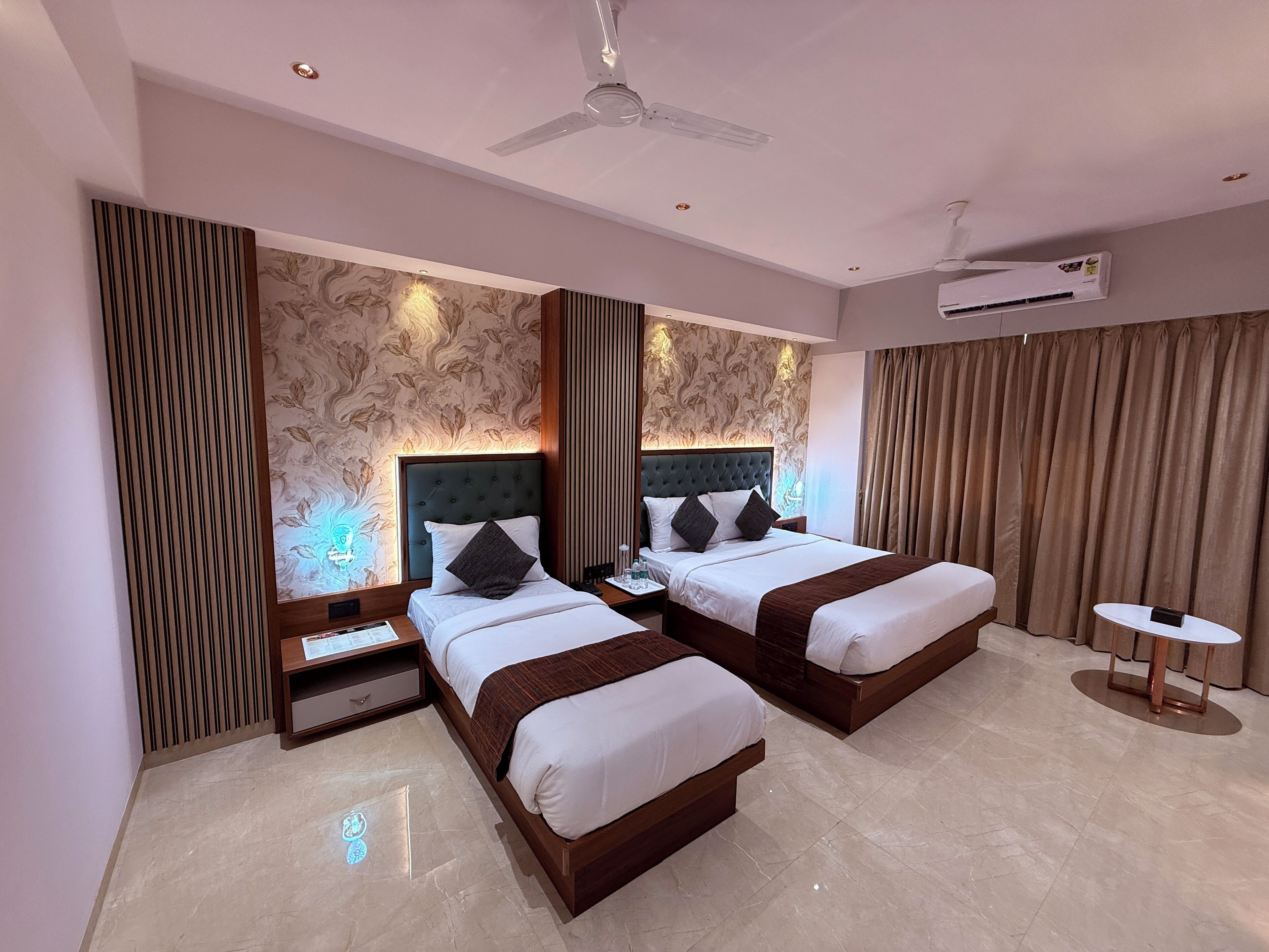 A hotel room with two beds, a ceiling fan, an air conditioning unit, a small table, and a decorative wall panel.