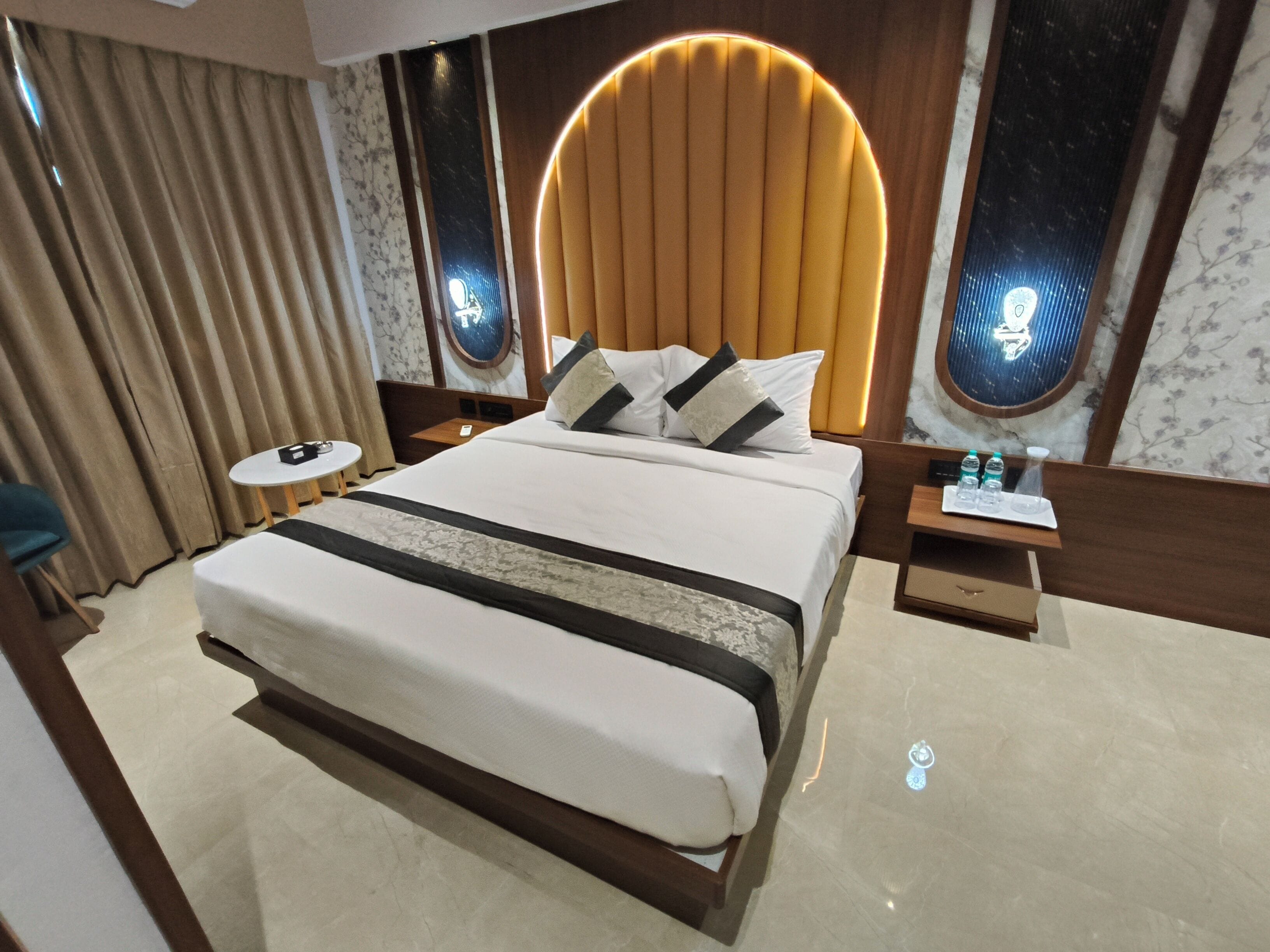 A modern hotel room with a large bed, a round headboard, a bedside table with a tray of water bottles, and a small round table.