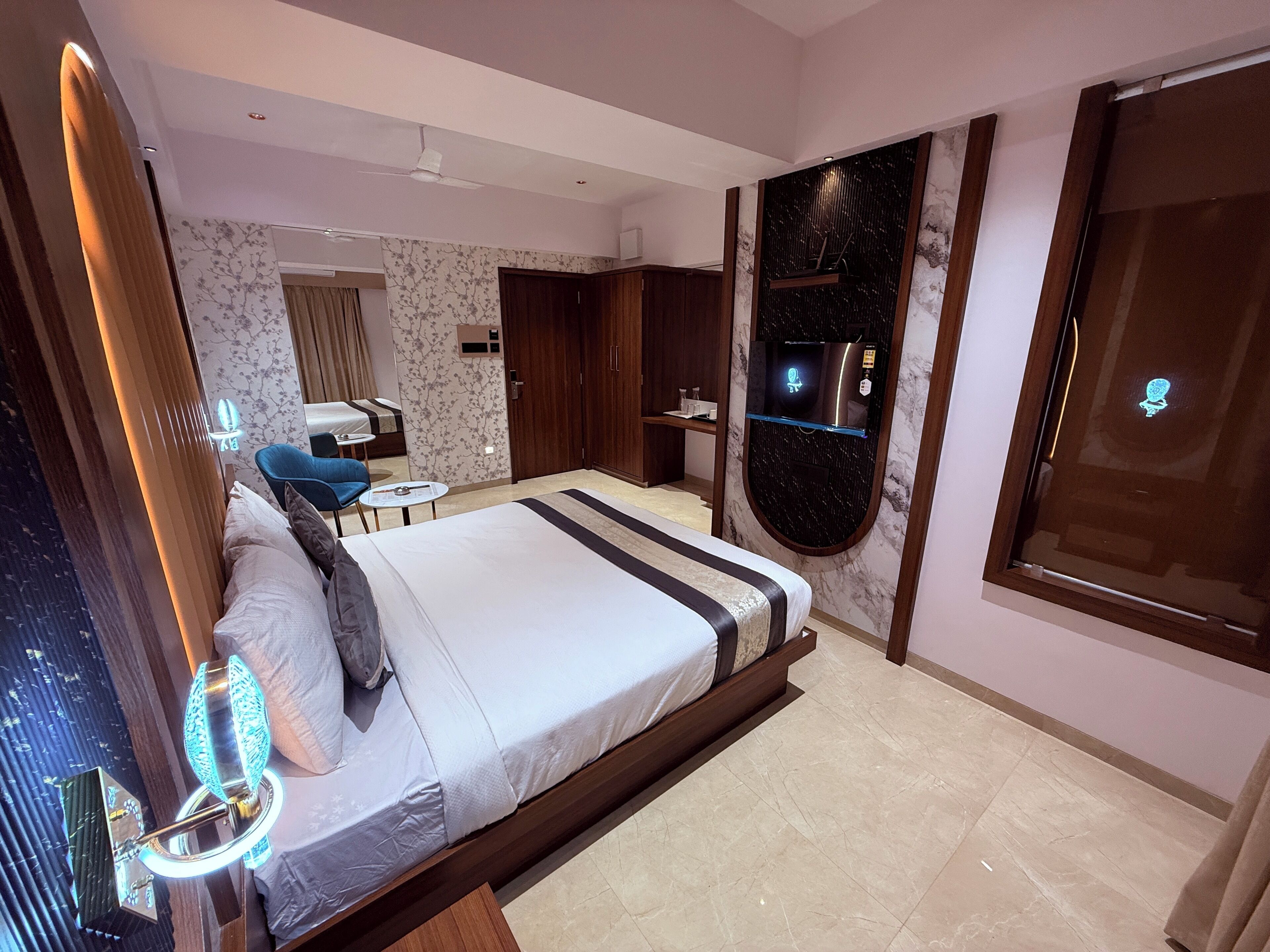 A modern hotel room with a large bed, a small table, a chair, a wardrobe, and a bathroom with a mirror and sink.