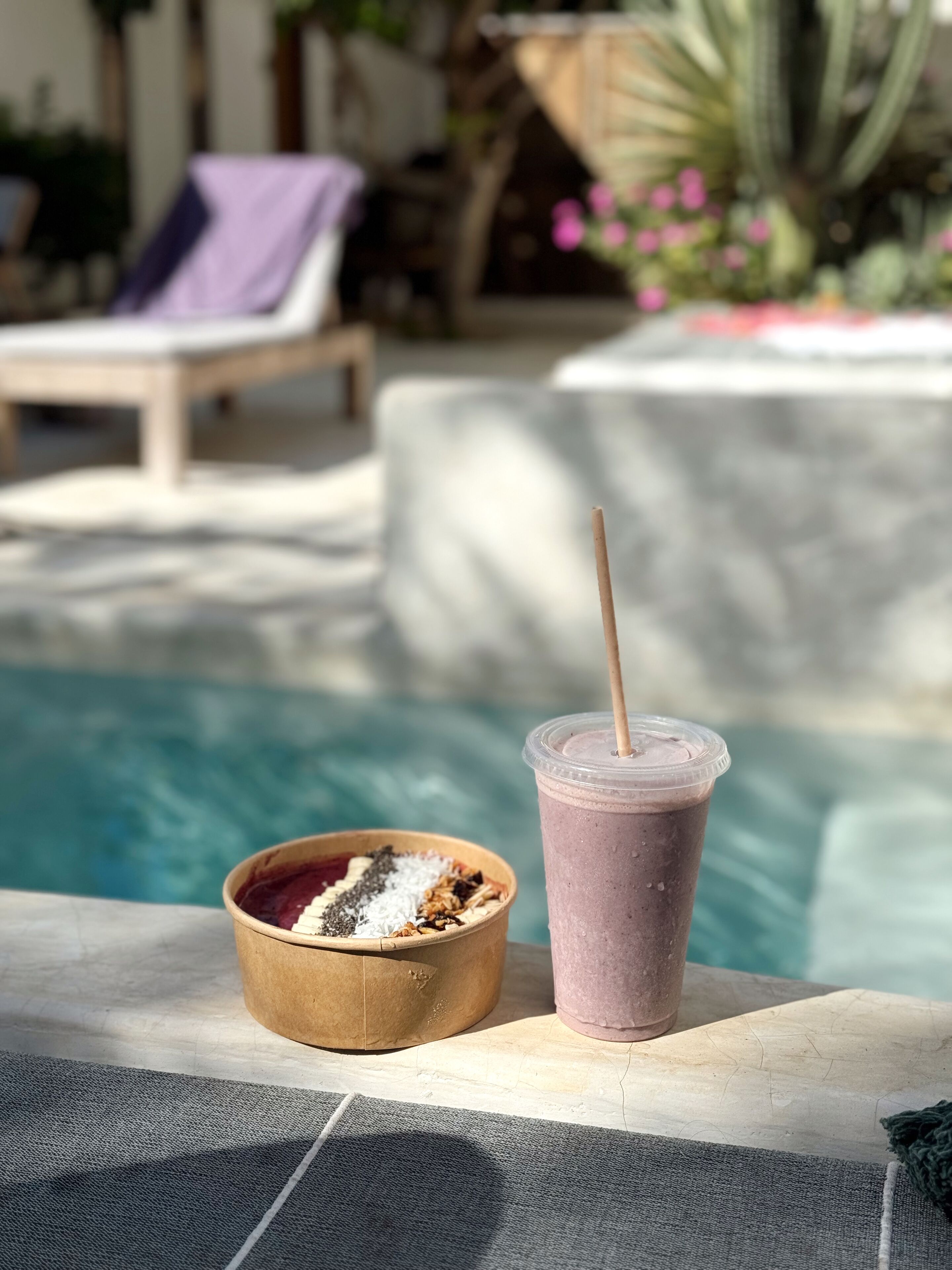 Escena junto a la piscina con una silla de madera, un plato de comida y un batido en vaso de plástico.