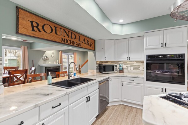 A modern kitchen with white cabinetry, a large wooden sign above the sink, and a view of the outside.