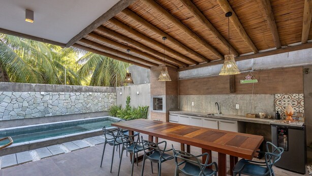An outdoor kitchen with a stone wall, a pool, and a wooden table with chairs. - House w/ Pool 140m From the Beach Clb0002 (São Miguel dos Milagres)