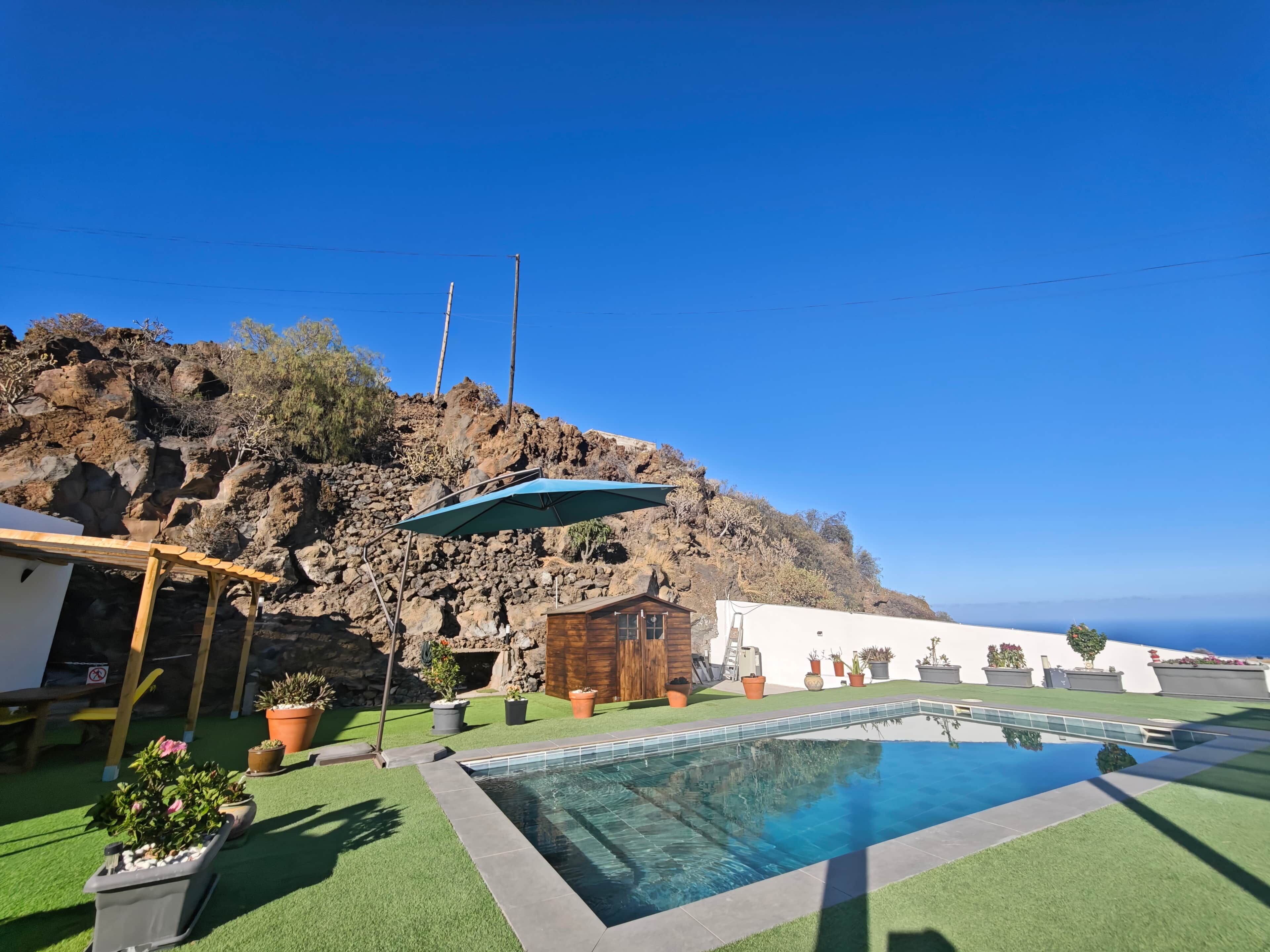 Un espace piscine avec un ciel bleu dégagé, une colline rocheuse et des plantes en pot.