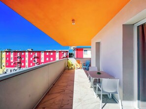 En balkon med flisegulv, et bord med stole og udsigt til farverige bygninger i baggrunden.