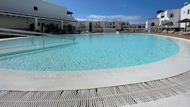 A large, clear swimming pool with a tiled deck, surrounded by white buildings with balconies and a clear blue sky. - The Balcony of the 4 Moons - Apartment 631 (Costa Teguise)
