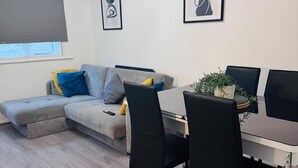Ένα μοντέρνο σαλόνι με έναν γκρι γωνιακό καναπέ, ένα τραπέζι τραπεζαρίας με καρέκλες και δύο έργα τέχνης σε κορνίζες στον τοίχο.