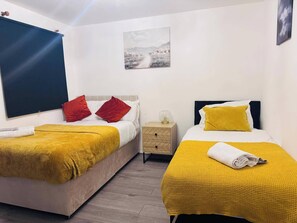 Habitación de hotel con dos camas, cada una con una manta amarilla y almohadas rojas. Hay una mesita de noche con una lámpara y una pequeña mesa con superficie de vidrio. Un cuadro cuelga en la pared sobre las camas.