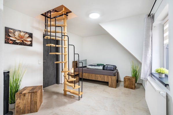 A loft bedroom with a wooden staircase, a bed with a gray mattress, and a window with a curtain.