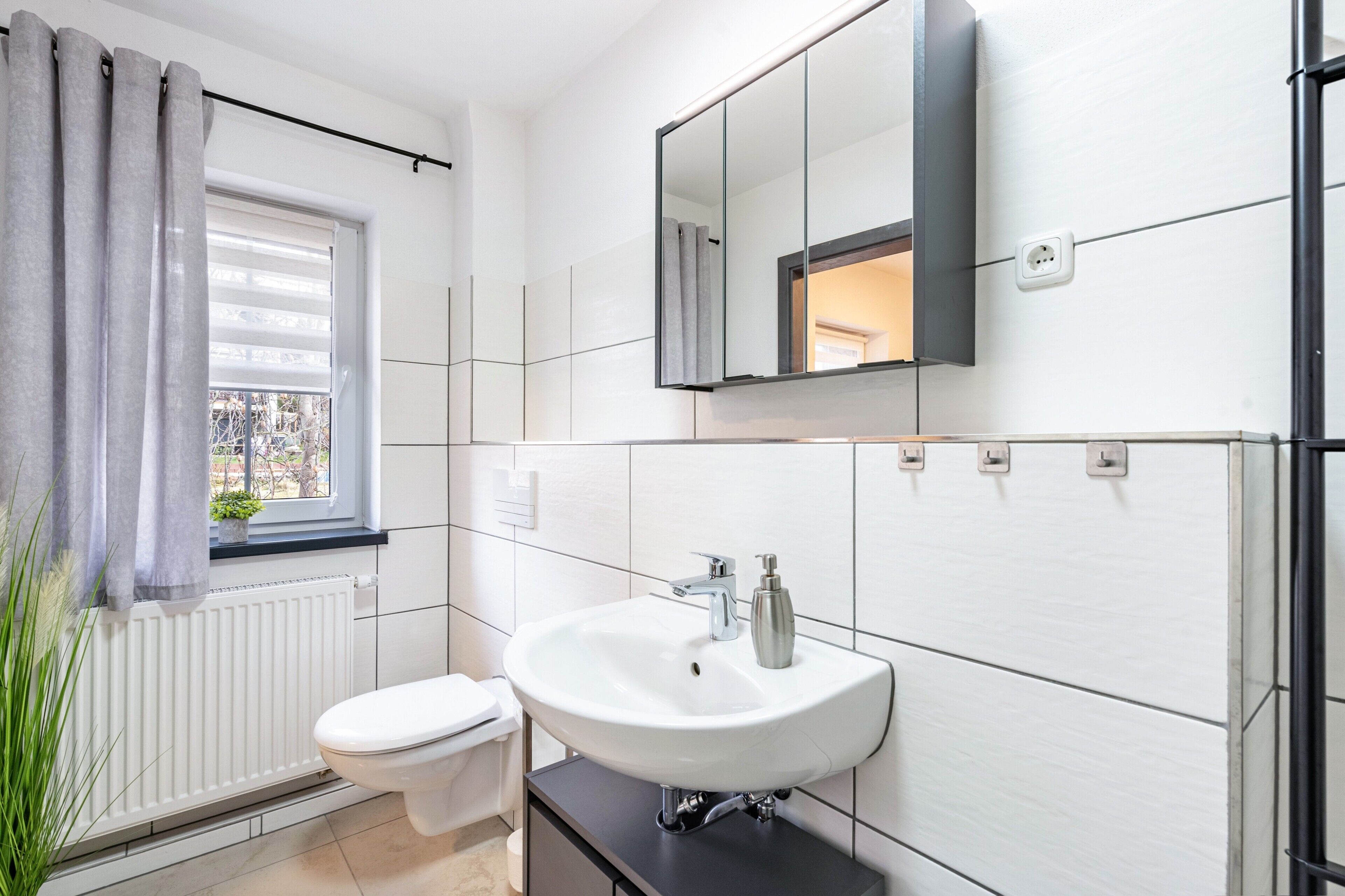 Une salle de bain avec des toilettes blanches, un lavabo avec un miroir au-dessus et une fenêtre avec des rideaux.