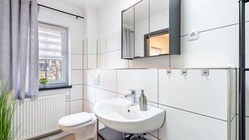 Une salle de bain avec des toilettes blanches, un lavabo avec un miroir au-dessus et une fenêtre avec des rideaux.