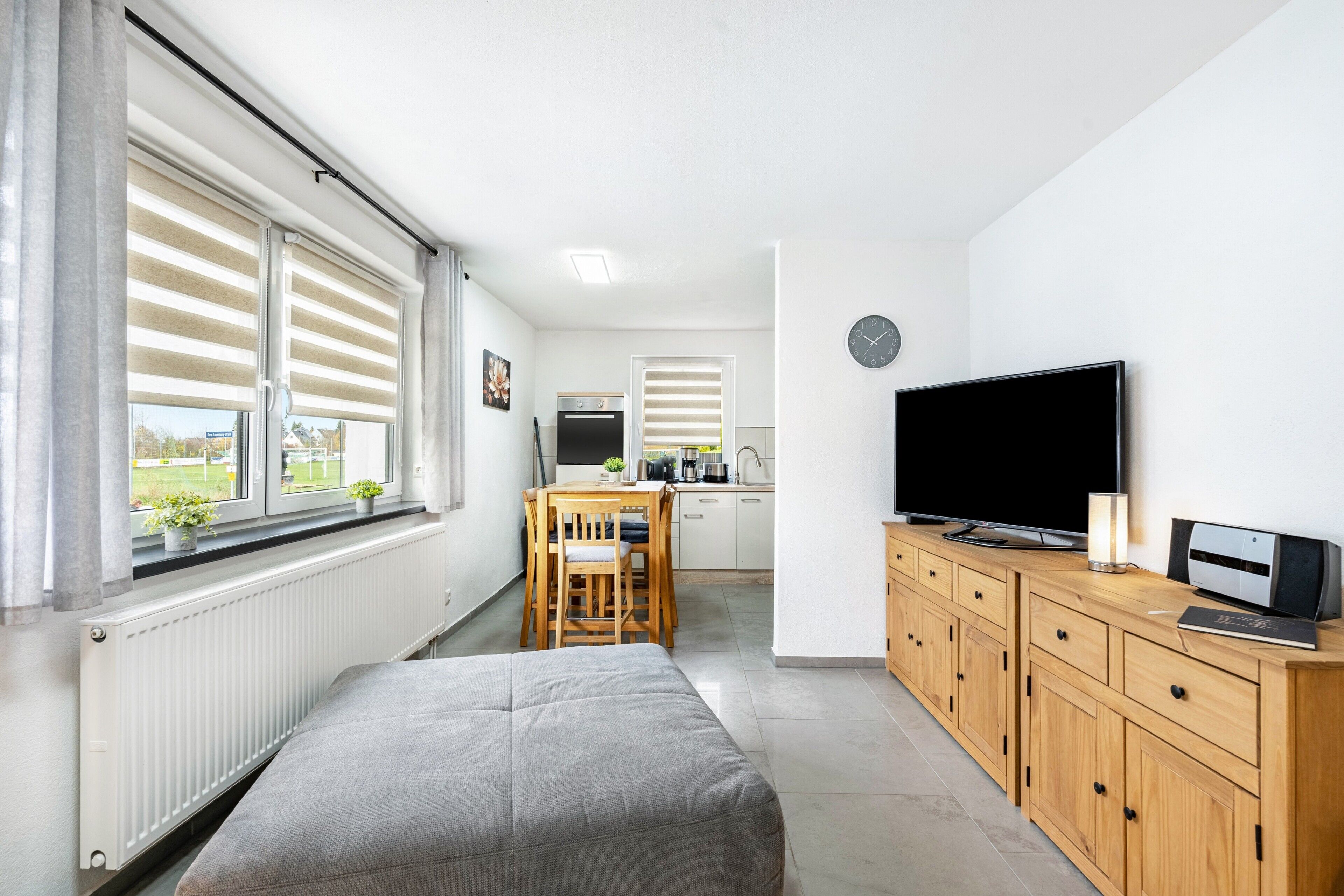 A modern room with a bed, a TV, a wooden cabinet, a kitchen area, and large windows with blinds.
