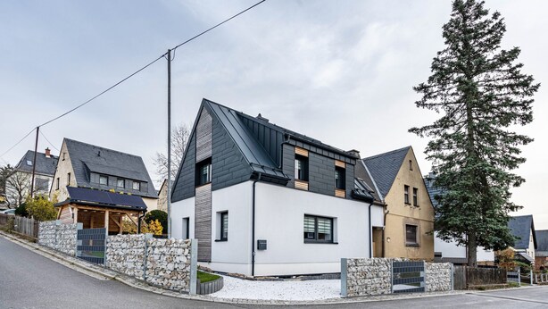 Et moderne hus med to etager, skråtag, stenvægge og et stort træ i baggrunden.