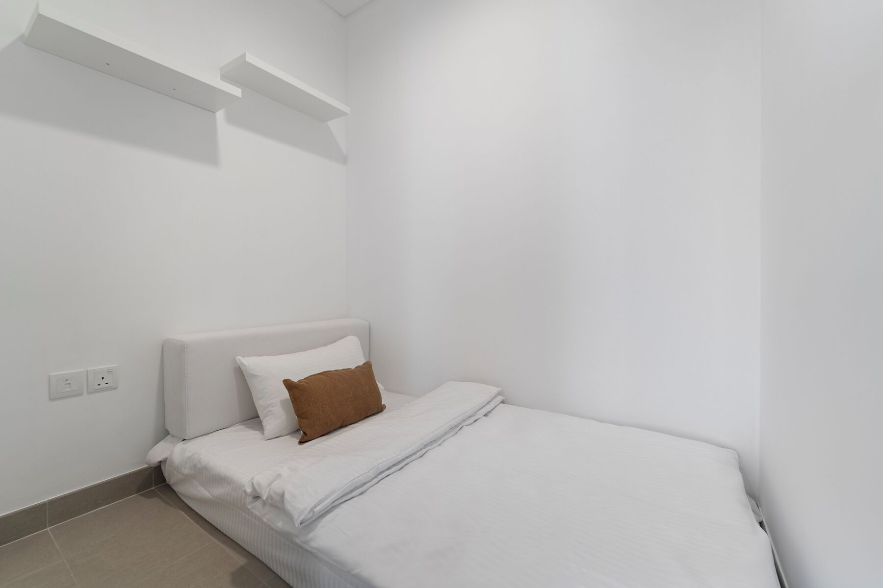 A small, white, single-bed room with a headboard, a brown pillow, and a white blanket.