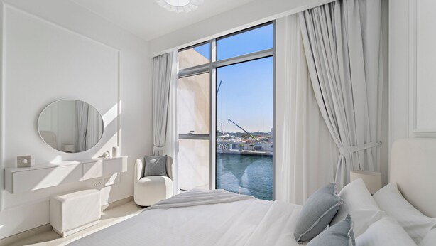 A modern bedroom with a large bed, a bedside table, a chair, a round mirror, and a view of a body of water through large windows. - 2BR M Room CF7 B Partial Canal View (Dubai)