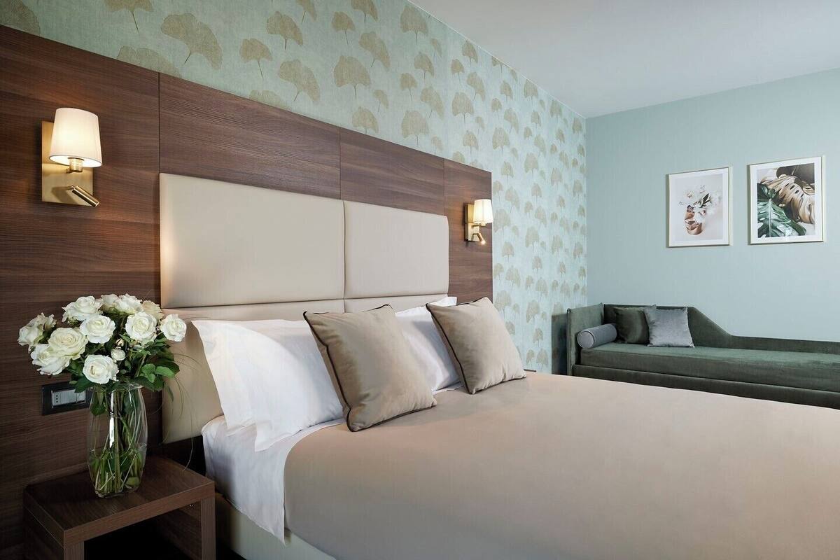 A hotel room with a large bed, a nightstand with a vase of flowers, a green sofa, and wall-mounted lamps.
