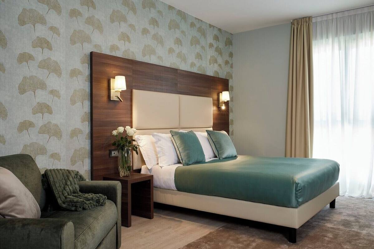 A hotel room with a large bed, a nightstand with a lamp, a green armchair, and a wall with a leaf pattern.
