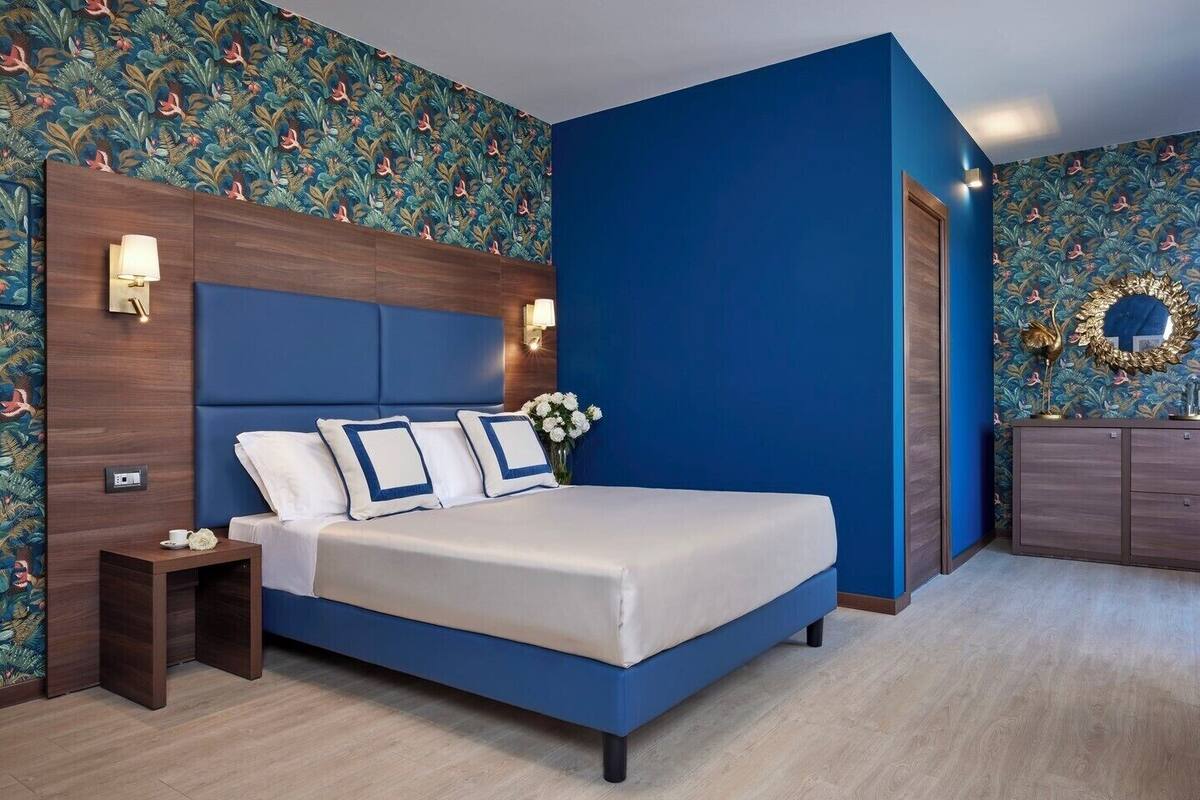 A modern hotel room with a large bed, a nightstand, a dresser, and a decorative wall with a floral pattern.
