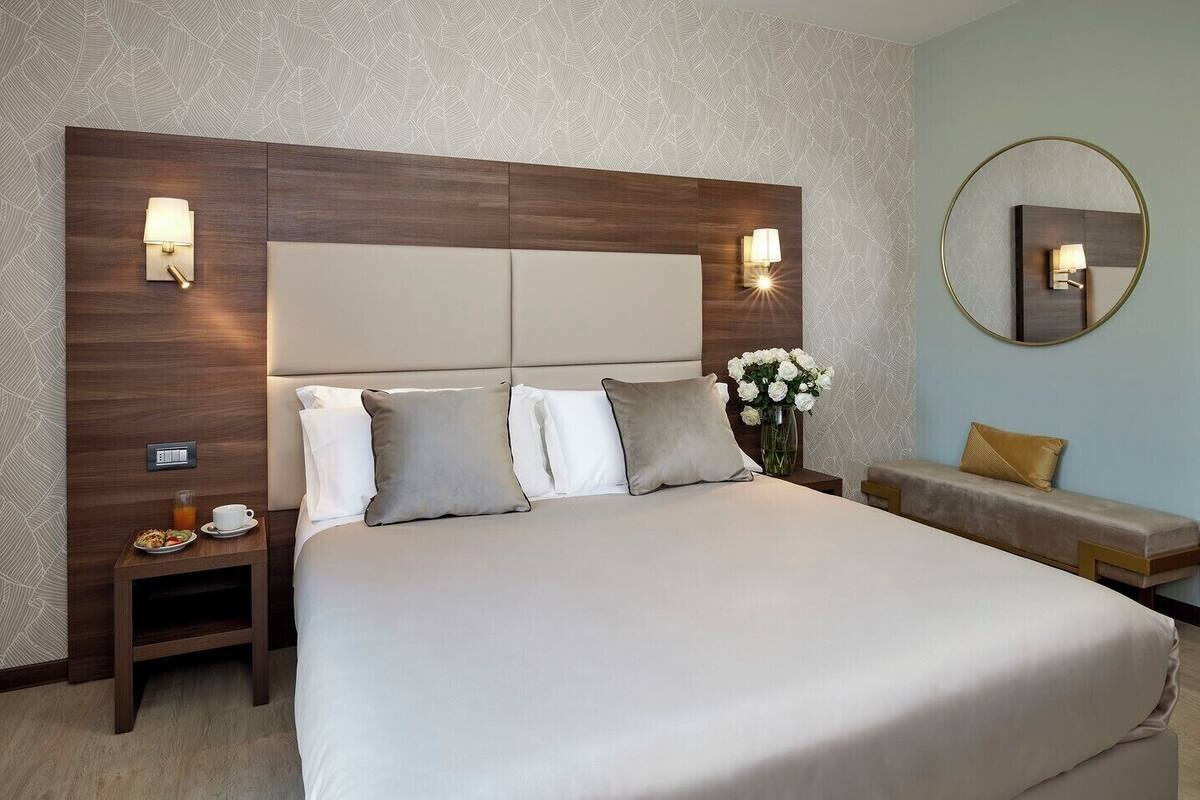 A hotel room with a large bed, two bedside tables, a nightstand, a vase with flowers, a round mirror, and a bench.