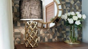 A room with a geometric patterned wall, a round mirror with a gold frame, a modern lamp with a textured shade, and a vase with white flowers on a wooden cabinet. - HOTEL SPIGAMONTI (Negrar)