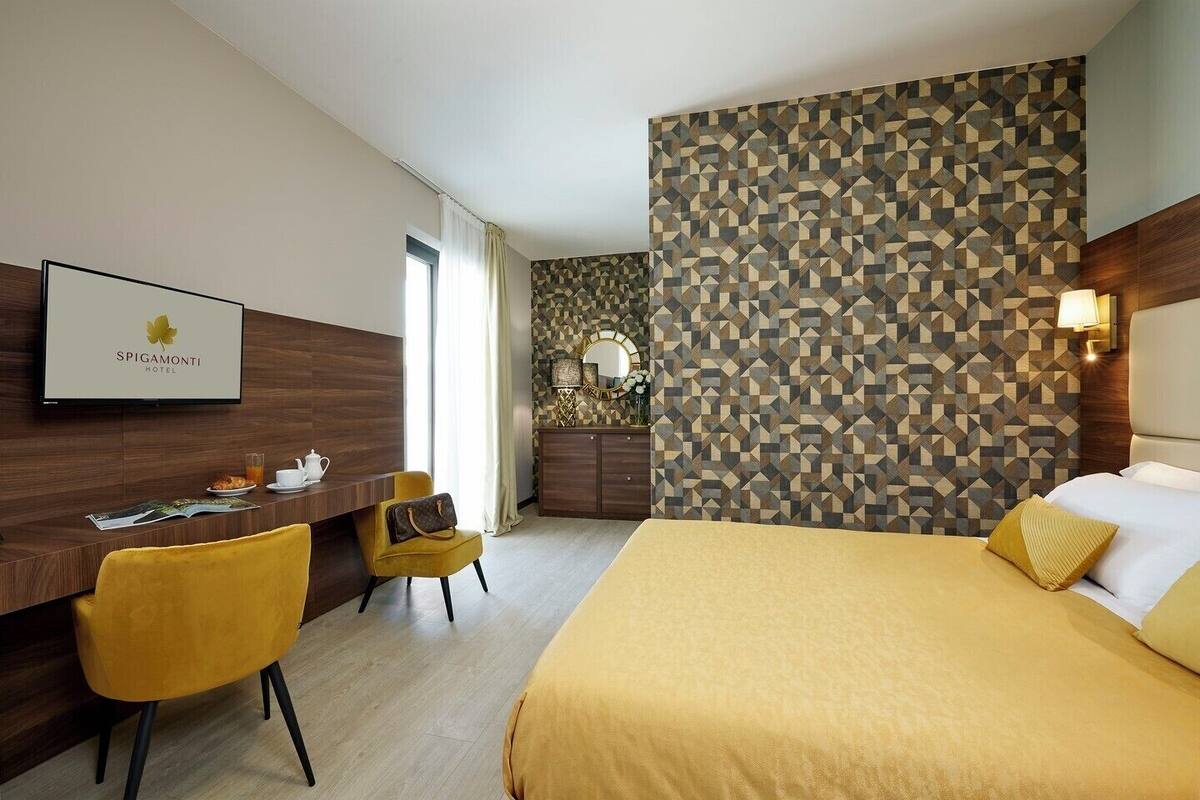 A hotel room with a bed, a desk with a chair, a TV, and a patterned wall.