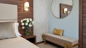 A hotel room with a bed, a nightstand with a vase of white flowers, a bench with a gold cushion, and a round mirror on the wall. - HOTEL SPIGAMONTI (Negrar)
