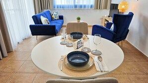 A dining area with a round table set for two, featuring a black bowl, wine glasses, and cutlery. Blue armchairs surround the table, and a sofa is visible in the background. - Stylish 1BR Apt. Pool Views | Private Patio | 1min walk to 5-star Gym (Pafos)