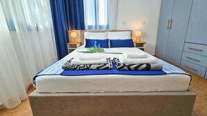 A neatly made bed with multiple pillows and a blanket, two bedside lamps, a small table, and a wardrobe with drawers. - Stylish 1BR Apt. Pool Views | Private Patio | 1min walk to 5-star Gym (Pafos)