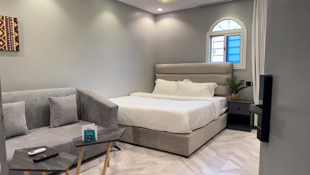 Un dormitorio moderno con un sofá gris, una mesita con un control remoto, una cama con ropa blanca y una ventana con cortinas.