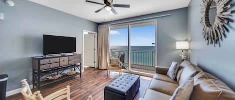 A living room with a flat-screen TV, a ceiling fan, a sofa, a chair, and a large window with a view of the ocean.