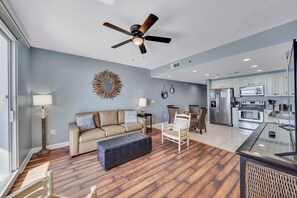 A spacious living area with a sofa, ottoman, and a dining table. A ceiling fan is visible, and the kitchen area is equipped with modern appliances. - NEW Listing! FREE Daily Golf&Perks! Splash W502 w/Hot Tub & Heated Pool &Gym! (Panama City Beach)