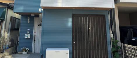 Un edificio residencial de varios pisos con puerta de garaje, un pequeño equipo de aire acondicionado y una planta en maceta cerca de la entrada.