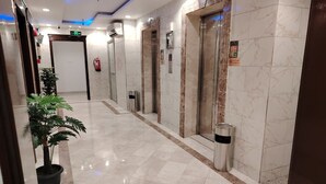 Een hotelhal met een marmeren vloer, liften, een potplant en een brandblusser.