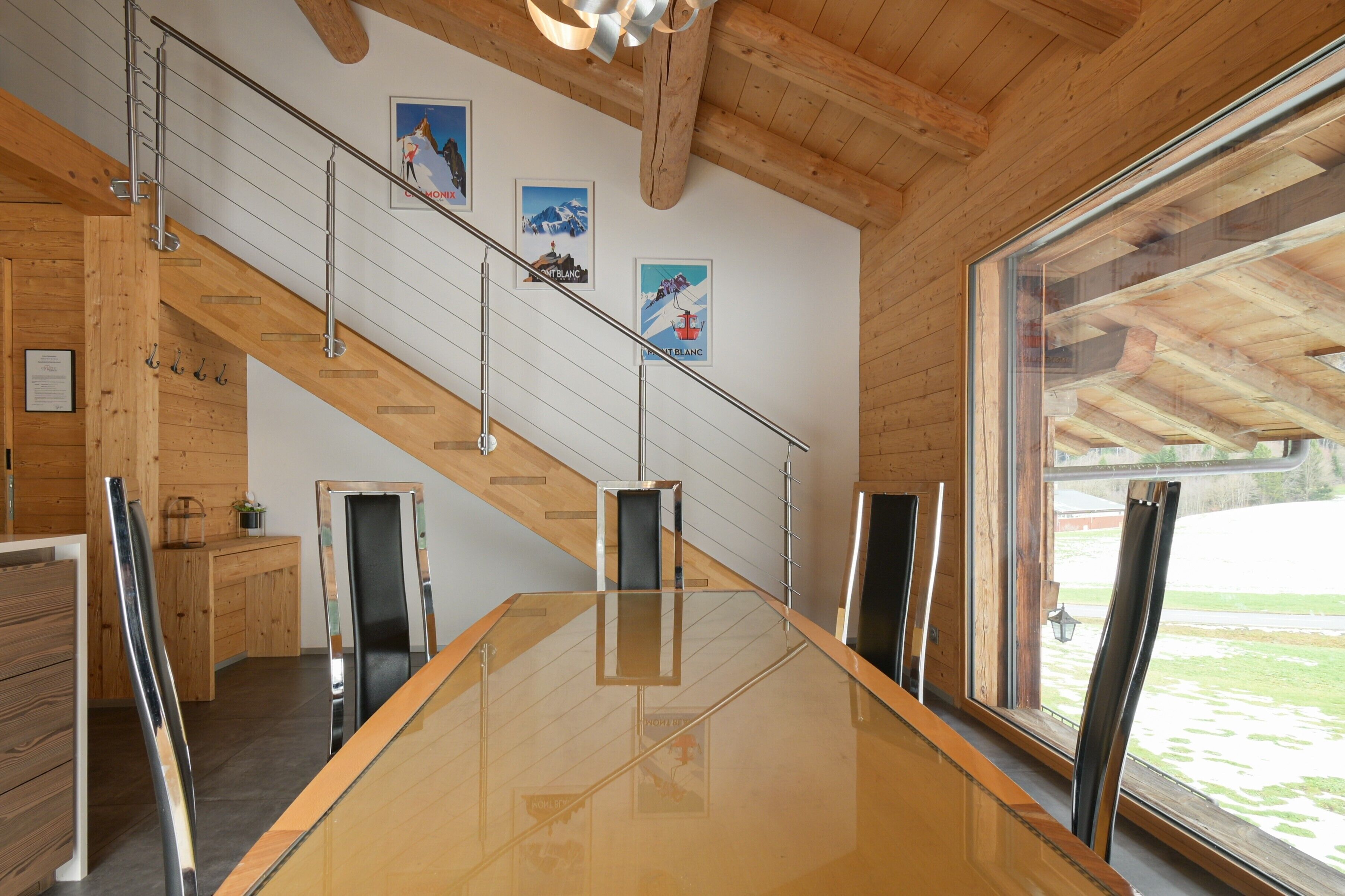 Un intérieur moderne avec un escalier en bois, une grande table en verre et une vue dégagée sur l’extérieur grâce à une grande fenêtre.