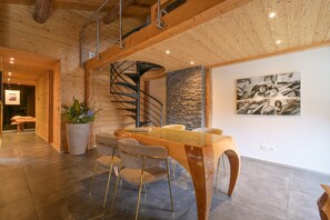 Ein modernes Interieur mit Holzbalkendecke, einer Wendeltreppe, einem Essbereich mit Glastisch und einer Wand mit Steinverkleidung.