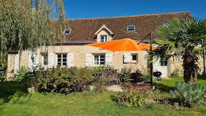Uma casa de pedra com telhado em telhas, um grande guarda-sol laranja e um jardim bem cuidado com várias plantas e uma palmeira.