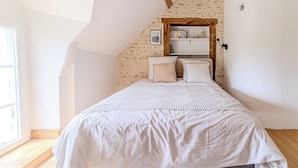 Quarto com teto inclinado, estante de madeira, cama com roupa de cama branca e janela que proporciona luz natural.