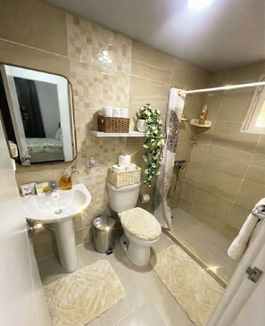 Et moderne badeværelse med vask, toilet og brusebad. Der er toiletartikler på hylderne, en skraldespand og et spejl.