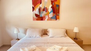 Cama bem arrumada com almofadas e um cobertor dobrado, uma luminária de cabeceira e um quadro abstrato colorido na parede.