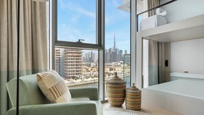 Quarto de hotel moderno com uma janela grande que oferece vista para a cidade, uma poltrona verde, dois vasos decorativos e uma mesa de cabeceira branca.
