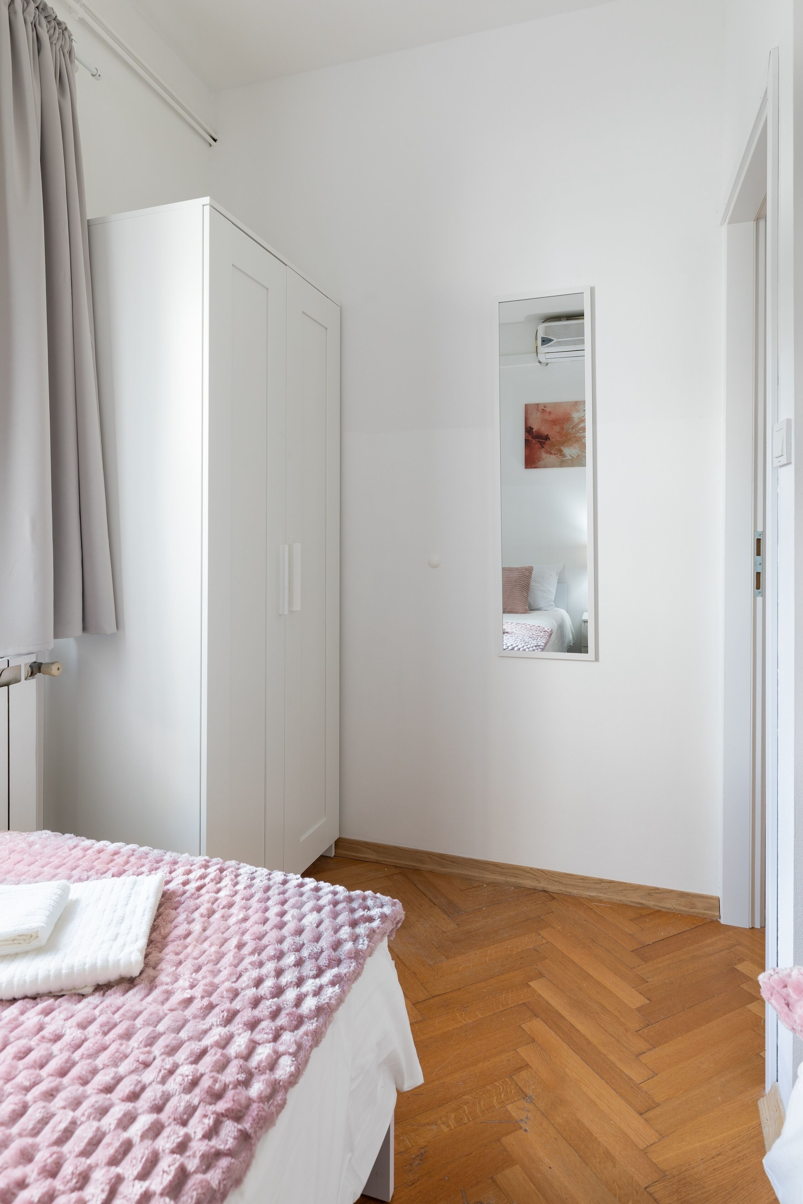 Ein Schlafzimmer mit Holzboden, einem Bett mit rosa strukturierter Decke, einem weißen Kleiderschrank und einem Spiegel an der Wand.