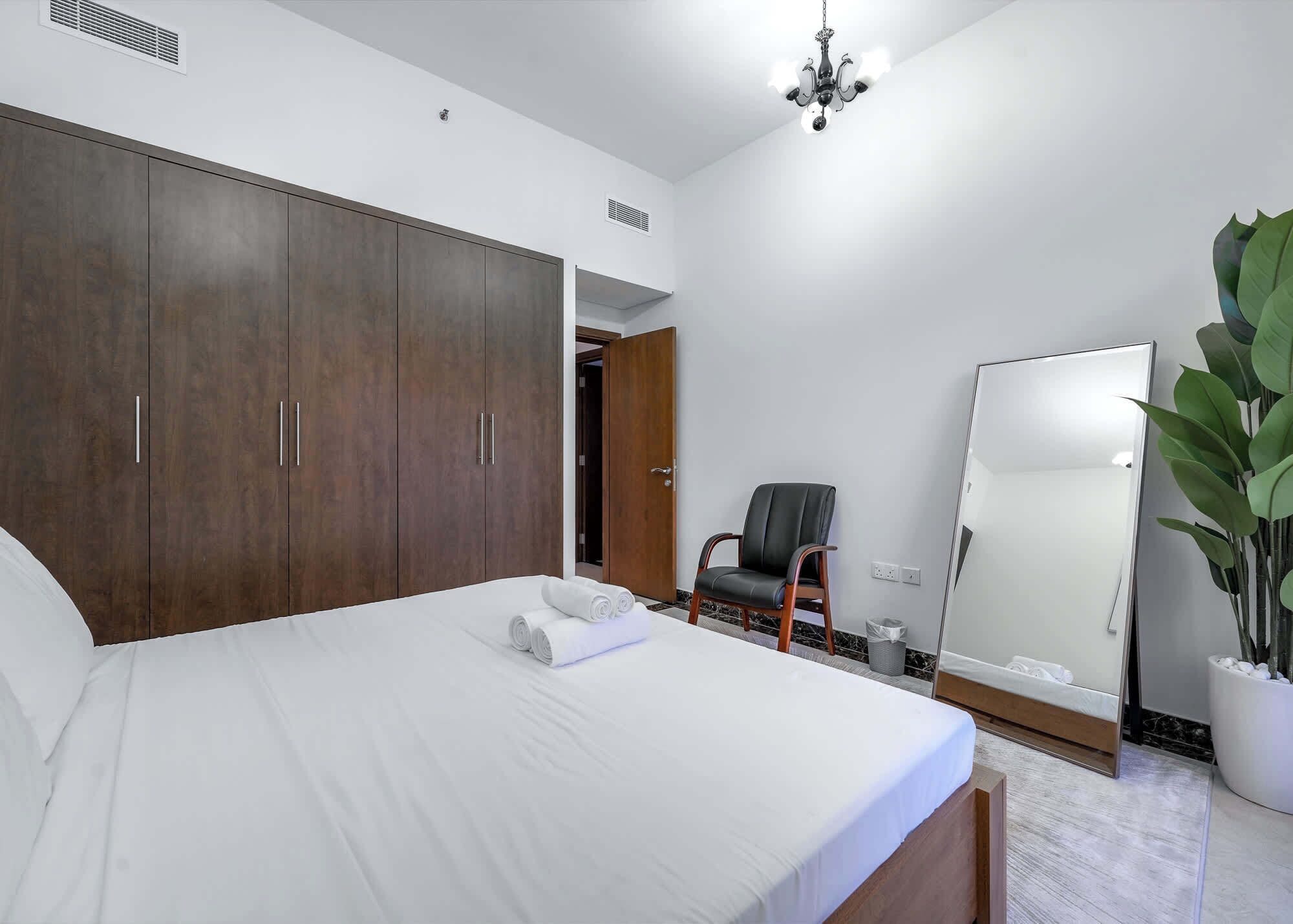 A bedroom with a large wardrobe, a bed with white linens, a chair, a mirror, and a potted plant.