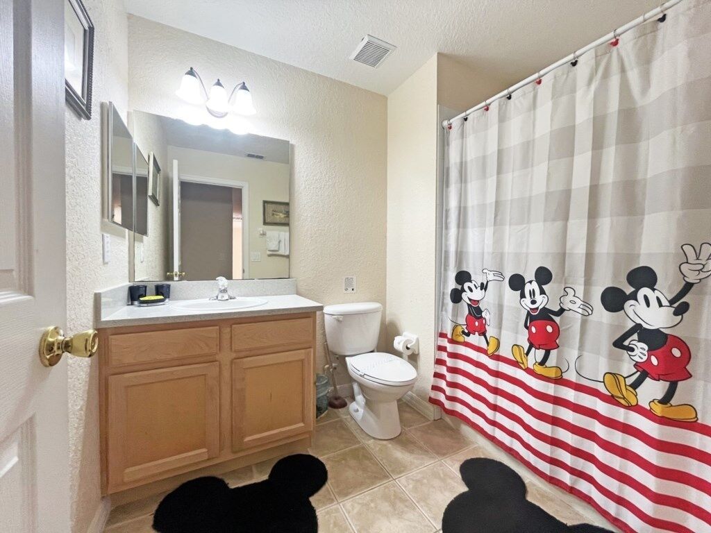 Une salle de bain avec des toilettes, un lavabo et un miroir. Il y a un rideau de douche à l’effigie de Mickey Mouse.