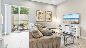 A modern living room with a sofa, coffee table, and TV. There are two framed artworks on the wall and a sliding glass door leading to an outdoor area. - Sunrise Shores 4 Bedroom Townhouse (Kissimmee)