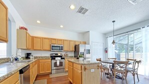 A modern kitchen with wooden cabinets, stainless steel appliances, and a dining area with a glass door to the outside. - Endless Summer 6 Bedroom Home (Kissimmee)