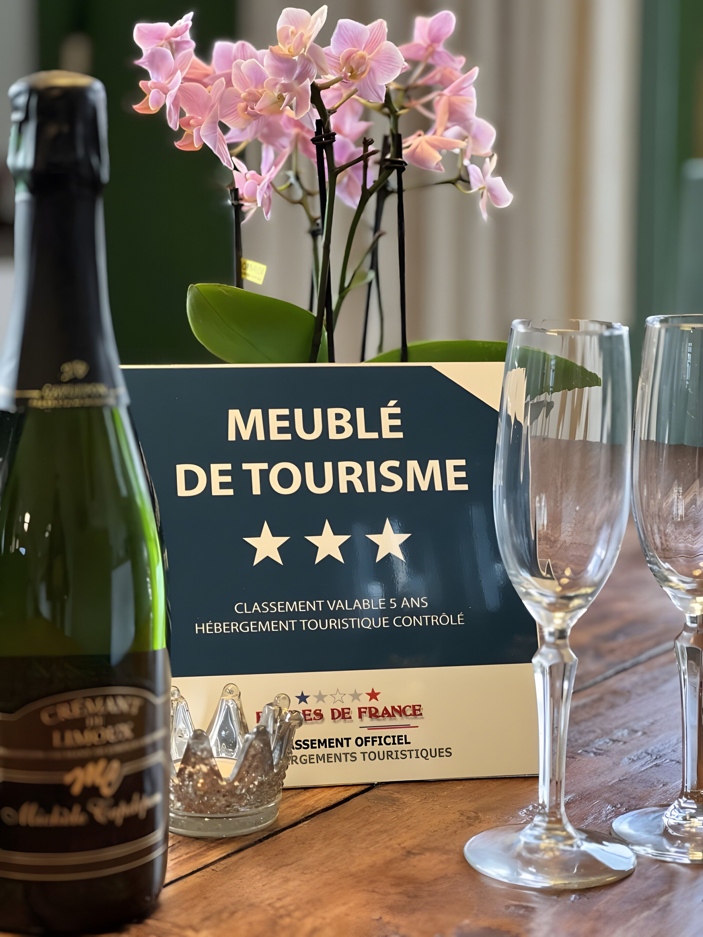 Een fles wijn, twee glazen, een doosje met de tekst 'MEUBLÉ DE TOURISME' en roze orchideeën.