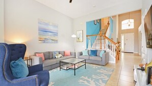 A spacious living room with a blue armchair, grey sofas, and a glass coffee table. A staircase leads to a second floor, and a painting adorns the wall. - Ohana Haven 5 Bedroom Home (Kissimmee)